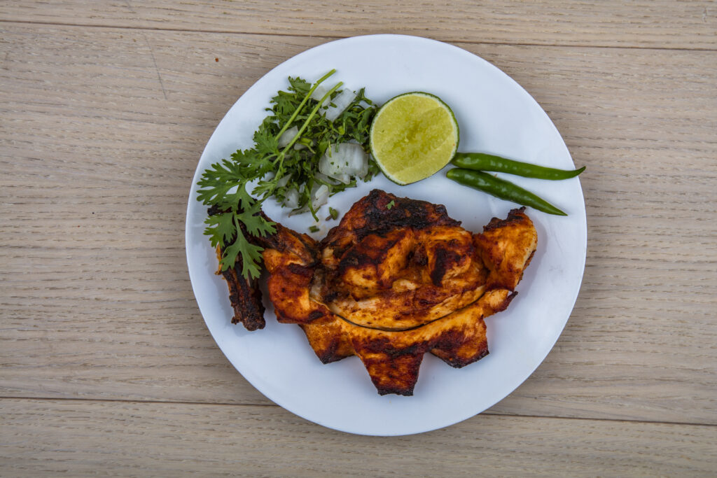 Grilled chicken tikka breast piece with distinct charred grill marks, marinated in rich spices, presented on a plate with a lemon wedge and fresh herbs for garnish.